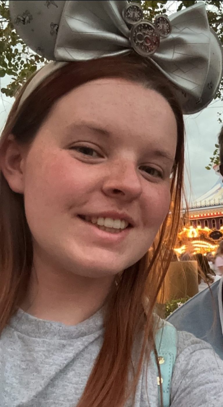 Young person with long red hair wearing sparkly silver mouse ears and a grey t-shirt.